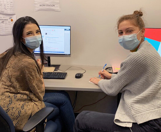 Yolanda Delmonte and Bella Shulman sitting around computer at desk with masks on.